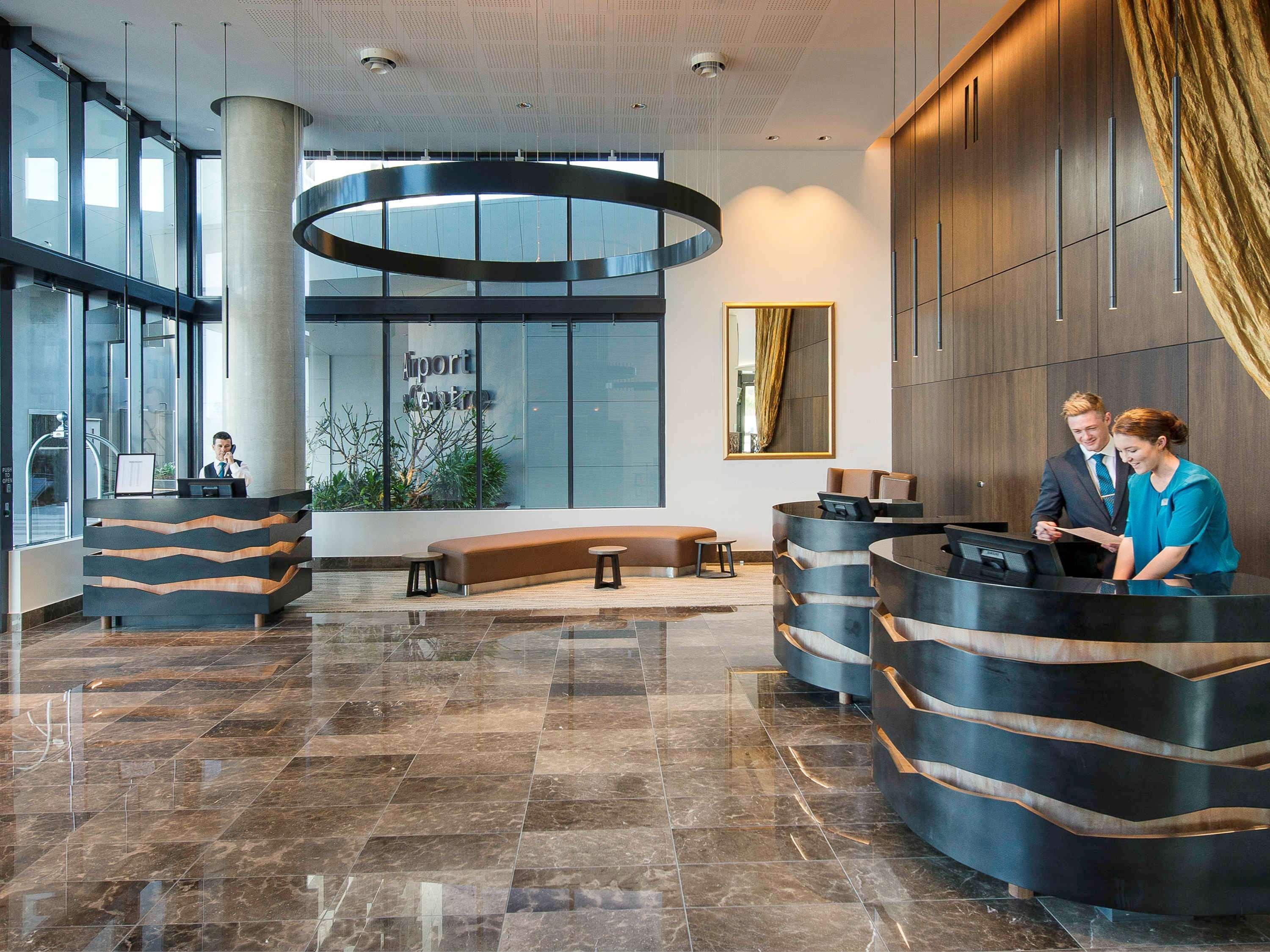 พูลแมน บริสเบน แอร์พอร์ต Hotel ภายนอก รูปภาพ The image shows a modern hotel lobby. There are two reception desks made of dark-colored materials, and staff members are attending to guests at these desks. The lobby features large windows that allow natural light to fill the space, and there are p